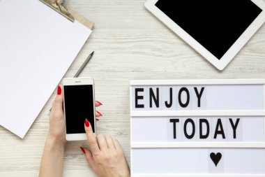 Tablet, smartphone, Not defteri ve lightbox 'Bugün Enjoy' kelime boşluk beyaz ahşap zemin, üstten görünüm üzerinde çalışıyorum. Eller telefon ile boş perde tutun. Yukarıdan, düz-lay, tepegöz. 