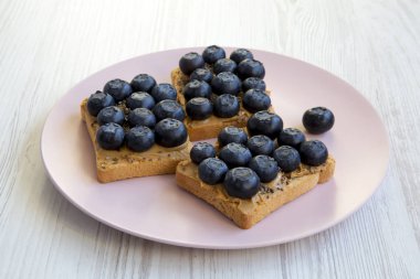 Fıstık ezmesi, yaban mersini ve beyaz ahşap yüzey, en iyi görünümü üzerinde pembe bir tabakta chia tohum ile vejetaryen tost. Sağlıklı beslenme. Yan görünüm.