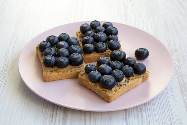 Fıstık ezmesi, yaban mersini ve beyaz ahşap yüzey, en iyi görünümü üzerinde pembe bir tabakta chia tohum ile vejetaryen tost. Sağlıklı beslenme. Yan görünüm.