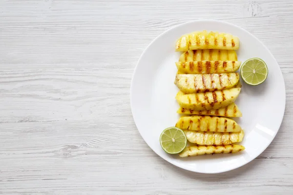 Grilled pineapple slices with lime on white plate and blank notebook over white wooden background, top view. Summer food. From above, overhead, flat lay. Copy space.