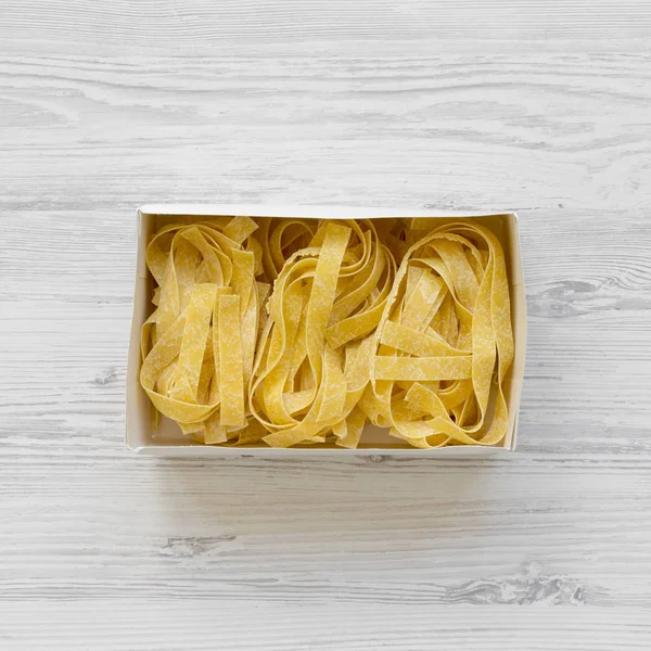 Uncooked pasta pappardelle in paper box on a white wooden background, view from above. Top view, overhead.