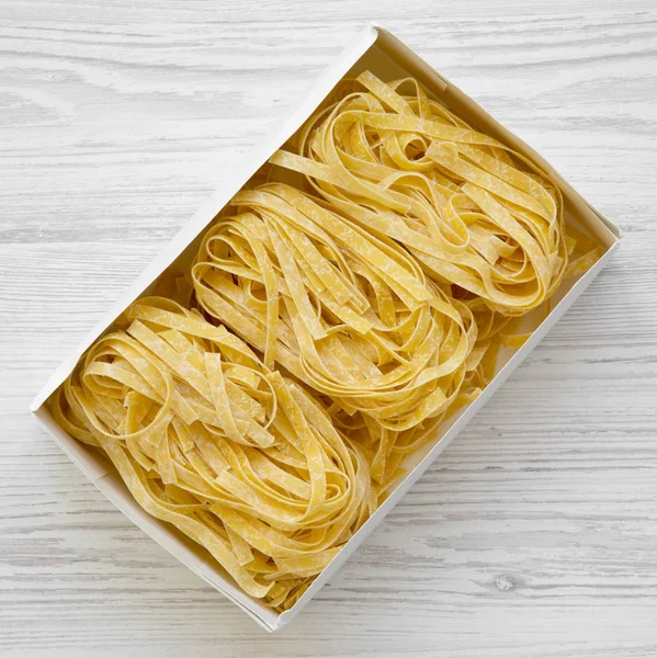 Uncooked pasta fettuccine in paper box on a white wooden background, top view. Overhead. Close-up.