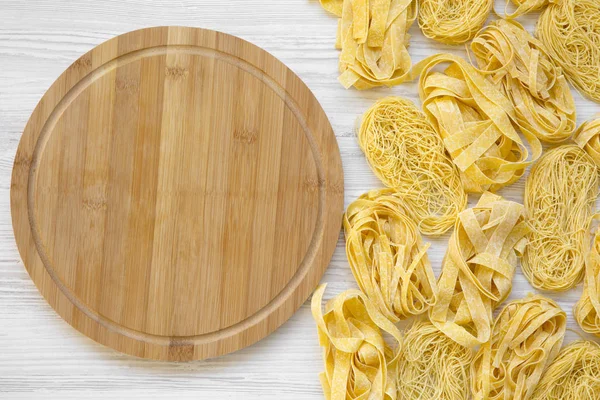 Various uncooked pasta with bamboo board on white wooden table, top view. From above, overhead, flat lay.