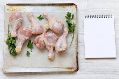 Çiğ çiğ tavuk ayakları üzerinde bir fırın yaprak kağıt tepsisi ve boş not defteri, havai görünümü. Beyaz ahşap arka plan. Metin için yer.