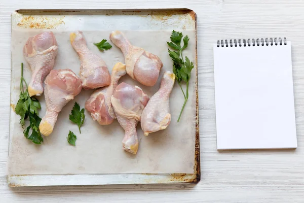 Çiğ çiğ tavuk ayakları üzerinde bir fırın yaprak kağıt tepsisi ve boş not defteri, havai görünümü. Beyaz ahşap arka plan. Metin için yer.
