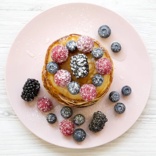 Pancakes with berries, honey and sugar on a pink plate on a white wooden table, overhead view. Flat lay, top view, from above. Close-up.