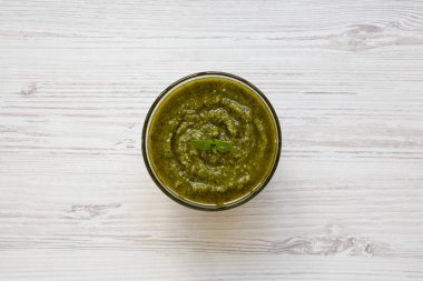 Beyaz ahşap arka planda, üstten görünüm taze yapılmış fesleğen pesto kase. Yukarıda, ek yükü.