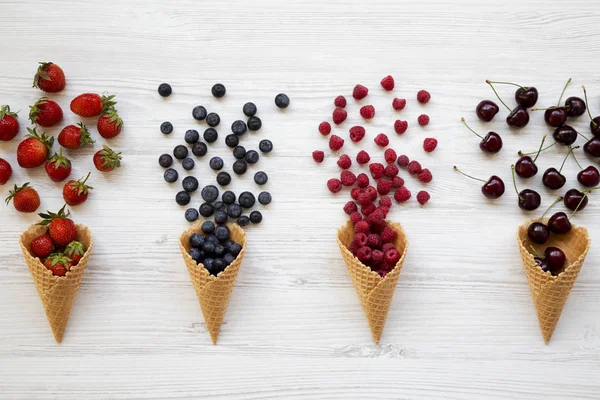 Waffle sweet ice cream cones with raspberries, cherries, strawberries and blueberries over white wooden background, overhead view. From above, top view.