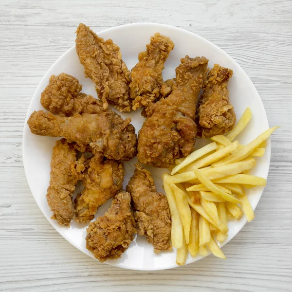 Tasty fried chicken drumsticks, spicy wings, French fries and chicken fingers on white plate over white wooden surface, overhead view. Flat lay, top view, from above.