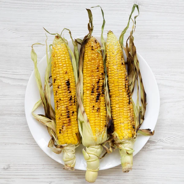 Grilled sweet corn on a white round plate over white wooden surface, top view. Summer vegan snack. Healthy diet. Close-up.