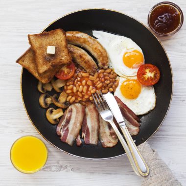 Tam İngiliz kahvaltısı bir tavada kızarmış yumurta, pastırma, sosis, fasulye, tost ve portakal suyu Beyaz ahşap yüzey, en iyi görünümü ile. Düz lay, ek yükü. Yukarıdan. 