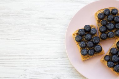 Üstten görünüm, vegan fıstık ezmesi, yaban mersini ve chia tohum pembe bir plaka üzerinde beyaz ahşap arka plan üzerinde kızartır. Yukarıdan Lay, düz. Kopya alanı.