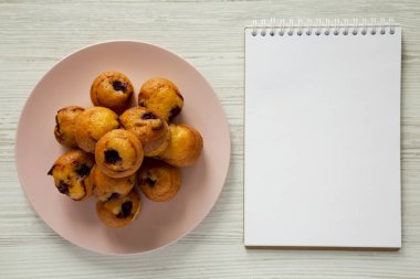 Üst görüntüleyin, kiraz pembe plaka üzerinde beyaz ahşap zemin üzerinde boş not defteri ile ev yapımı mini kekler. Tepegöz, yukarıdan, lay düz.