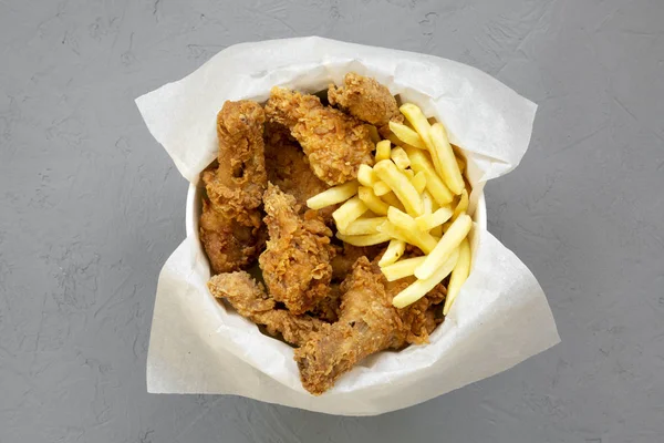 Tasty fast food: fried chicken drumsticks, spicy wings, French fries and chicken fingers in paper box over gray background, top view. Flat lay, from above, overhead.