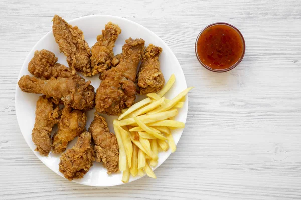 Tasty fried chicken drumsticks, spicy wings, French fries, chicken fingers and sauce on white plate over white wooden background, top view. Flat lay, overhead, from above.