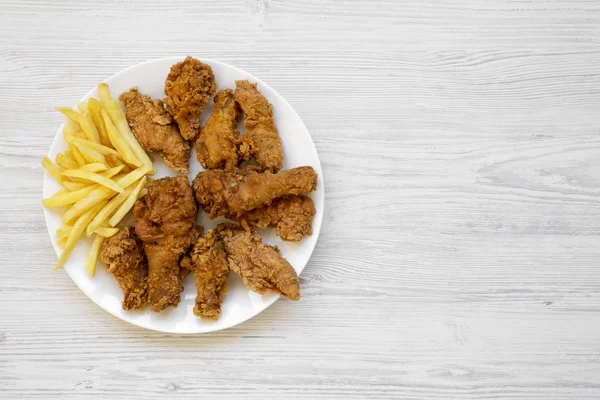 Tasty fried chicken legs, spicy wings, French fries and chicken fingers on white plate over white wooden background, top view. Flat lay, overhead, from above. Copy space.