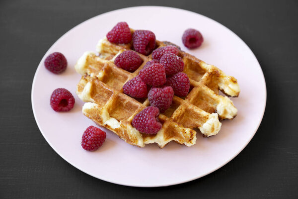 Traditional belgian waffle with raspberries on pink plate over black surface, side view. Close-up.