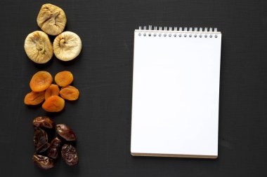 Siyah arka planda kurutulmuş meyveler ve boş not defteri, üst görünüm. 