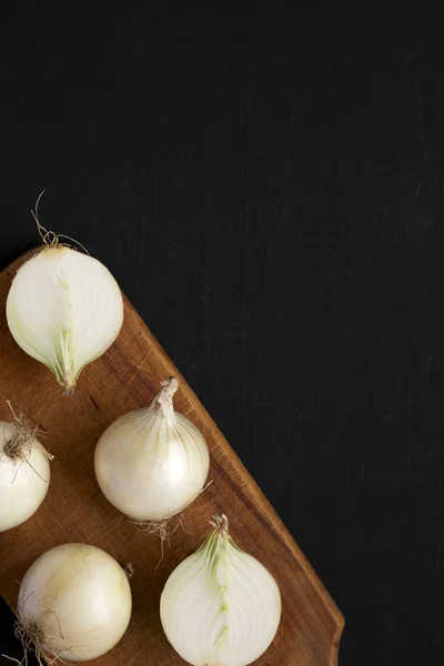 Raw white onions on a rustic board on a black surface, top view.