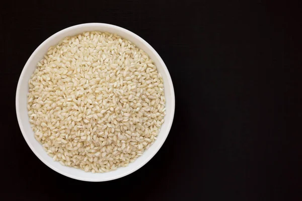 Organic Arborio rice in a white bowl on a black background, top — Stock Photo, Image