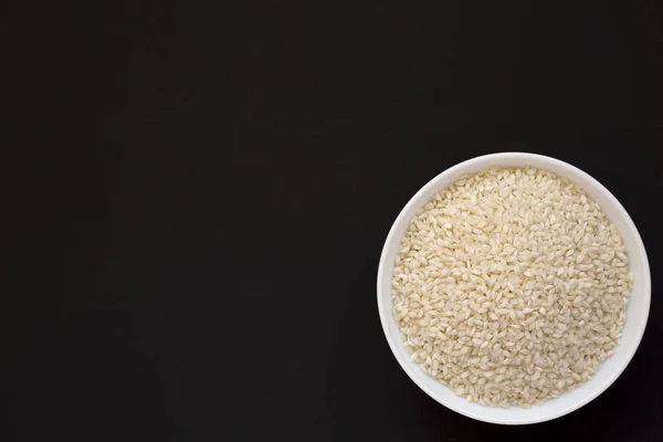 Organic Arborio rice in a white bowl on a black surface, top vie — Stock Photo, Image