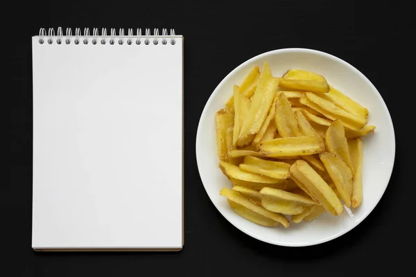 Zelfgemaakte frietjes op een wit bord, blanco notitieblok op een zwart — Stockfoto
