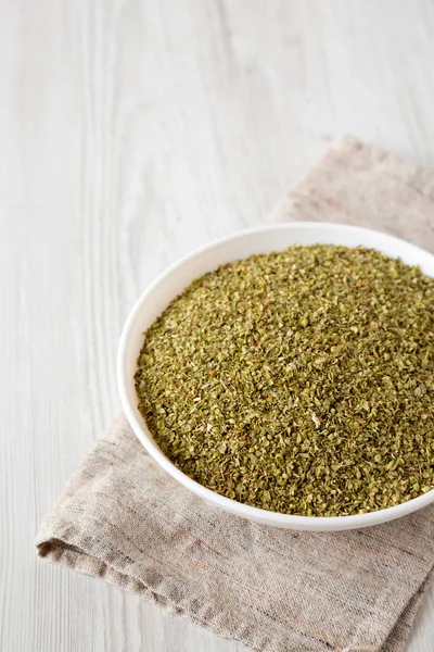 Dried Green Greek Oregano Spice in a white bowl on a white wooden surface, side view. Copy space.