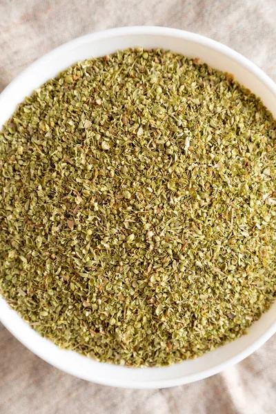 Dried Green Greek Oregano Spice in a white bowl on cloth, overhead view. Flat lay, from above, top view. Close-up.