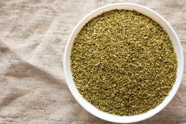 Dried Green Greek Oregano Spice in a white bowl on cloth, overhead view. Flat lay, top view, from above. Copy space.