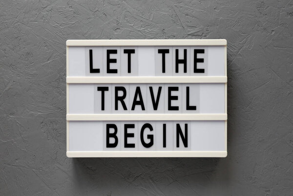 'Let the travel begin' on a lightbox on a gray background, top view. Flat lay, from above, overhead.