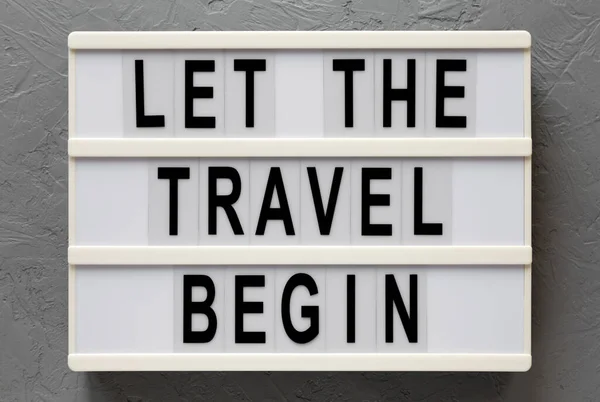 \'Let the travel begin\' on a lightbox on a gray surface, top view. Flat lay, from above, overhead. Close-up.