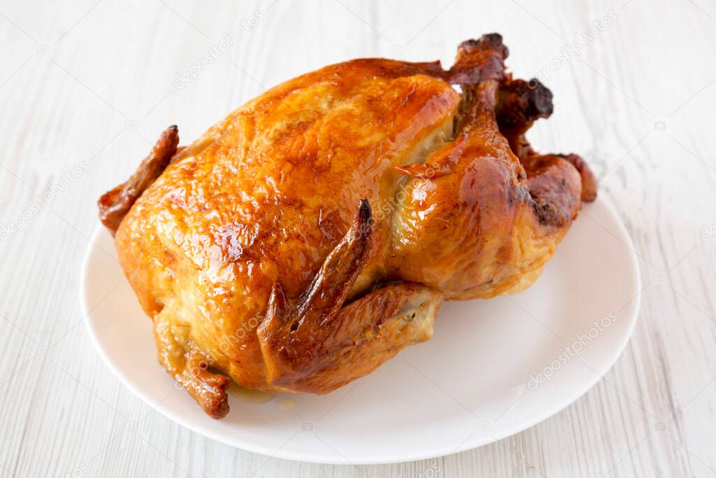 Homemade tasty rotisserie chicken on white plate on a white wooden background, side view. Close-up.