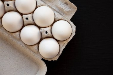 Siyah bir yüzey üzerinde, kağıt bir kutuda pişmemiş organik beyaz yumurta. Dümdüz, tepeden, yukarıdan. Boşluğu kopyala.