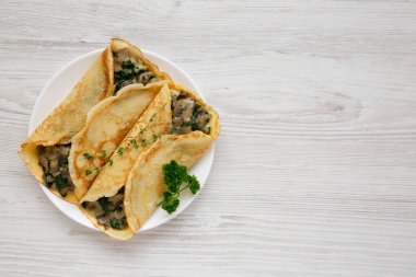 Beyaz ahşap bir yüzey üzerinde beyaz bir tabakta ev yapımı mantar, ıspanak ve peynir krepleri, üst manzara. Dümdüz, tepeden, yukarıdan. Boşluğu kopyala.