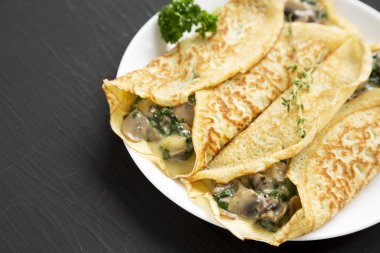 Beyaz bir tabakta siyah arka planda ev yapımı mantar, ıspanak ve peynirli krep. Boşluğu kopyala.