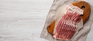 Beyaz ahşap arka planda, ahşap bir tahtanın üzerinde pişmemiş çiğ domuz pastırması. Düz yatıyordu, yukarıdan, yukarıdan. Metin için boşluk.