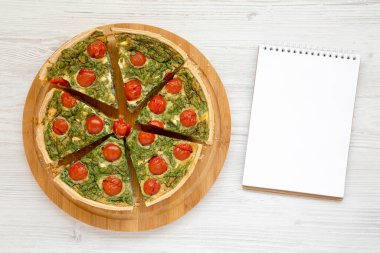 Yuvarlak bir bambu tahtada ev yapımı ıspanaklı kiş, beyaz ahşap bir yüzeyde boş not defteri, üst manzara. Dümdüz, tepeden, yukarıdan. 