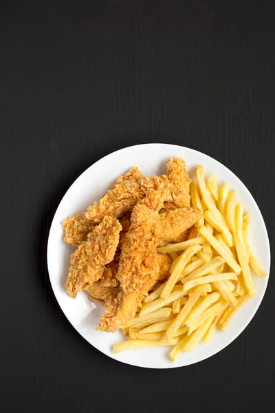 Homemade Crispy Chicken Tenders and French Fries on a white plate on a black surface, top view. Space for text.