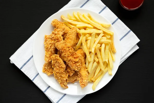 Homemade Crispy Chicken Tenders and French Fries on a white plate on a black surface, top view. Flat lay, overhead, from above.