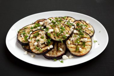 Siyah bir yüzey üzerinde beyaz bir tabakta Feta ve şifalı otlu ev yapımı ızgara patlıcan..