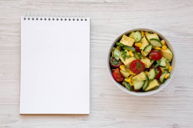 Kâsede taze Avocado Domates Salatası, beyaz ahşap arka planda boş not defteri, üst manzara. Dümdüz, tepeden, yukarıdan. 