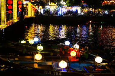 Fener yanan tekneler Hoi Nehri boyunca yüzer. Geceleri parlayan binalar ve hareketli kalabalıkla bir nehir. Hoi An, Vietnam, Temmuz 2025