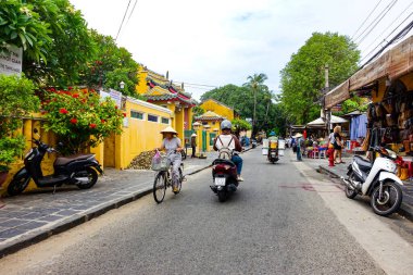 Eski Hoi An caddesinde Vietnamlı kadın bisikletleri, geleneksel Vietnam şapkalarıyla süslenmiş, zamansız bir sahne yaratıyor. Hoi An, Vietnam, Temmuz 202