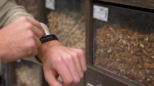 年轻人在柜台附近用坚果计算他的卡路里 手臂上有一个带有热量的健身跟踪器 盖伊通过手镯检查超市里的钻头的到期日期 — 图库视频影像