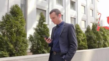 Genç işadamı sokakta yürüyor ve akıllı telefona bir mesaj yazıyor. Klasik takım elbiseli adam kendi telefonunu kullanıyor. Arka planda küçük yeşil ağaçlar var. 4k görüntü.