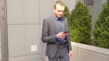 Genç işadamı sokakta yürüyor ve akıllı telefona bir mesaj yazıyor. Klasik takım elbiseli adam kendi telefonunu kullanıyor. Arka planda küçük yeşil ağaçlar var. 4k görüntü.