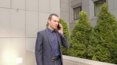 Genç bir işadamı sokakta yürüyor ve telefon görüşmesi hakkında konuşuyor. Klasik takım elbiseli bir adam akıllı telefonda bir konuşma yönetir. Arka planda küçük yeşil ağaçlar. 4k görüntü.