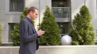 Genç işadamı sokakta yürüyor ve akıllı telefona bir mesaj yazıyor. Klasik takım elbiseli adam kendi telefonunu kullanıyor. Arka planda küçük yeşil ağaçlar var. 4k görüntü.