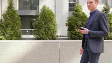 Genç bir işadamı sokakta yürürken kameraya bakarken gülümsüyor. Elinde bir akıllı telefon tutan klasik takım elbiseli adam. Backgroun'daki küçük yeşil ağaçlar. 4k görüntü.