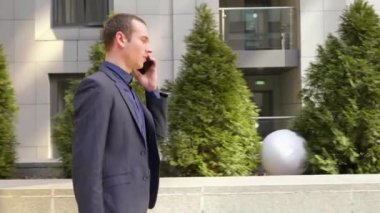 Genç bir işadamı sokakta yürüyor ve telefon görüşmesi hakkında konuşuyor. Klasik takım elbiseli bir adam akıllı telefonda bir konuşma yönetir. Arka planda küçük yeşil ağaçlar. 4k görüntü.